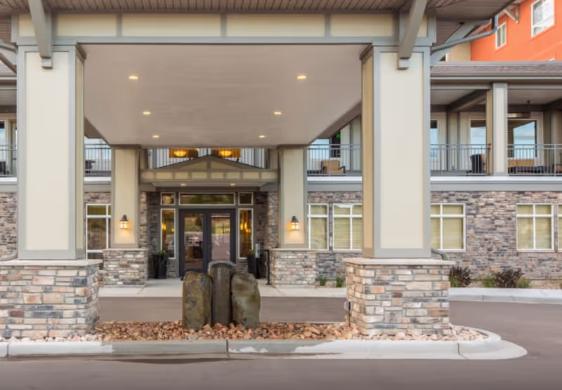 Entrance of a senior living facility with a covered drop-off area supported by large columns with stone bases. The building exterior features stone and beige paneling with windows and a balcony above the entrance. There are three large decorative rocks surrounded by small stones in the center of the driveway area.