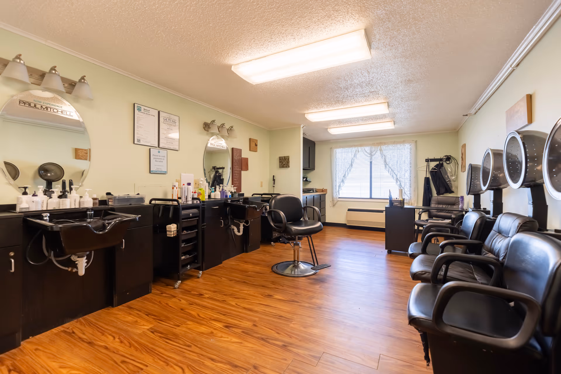 Interior view of a hair salon area with black salon chairs, hair washing sinks, mirrors, and hair dryers mounted on the wall. The room has wooden flooring, light-colored walls, and a window with lace curtains allowing natural light to enter.