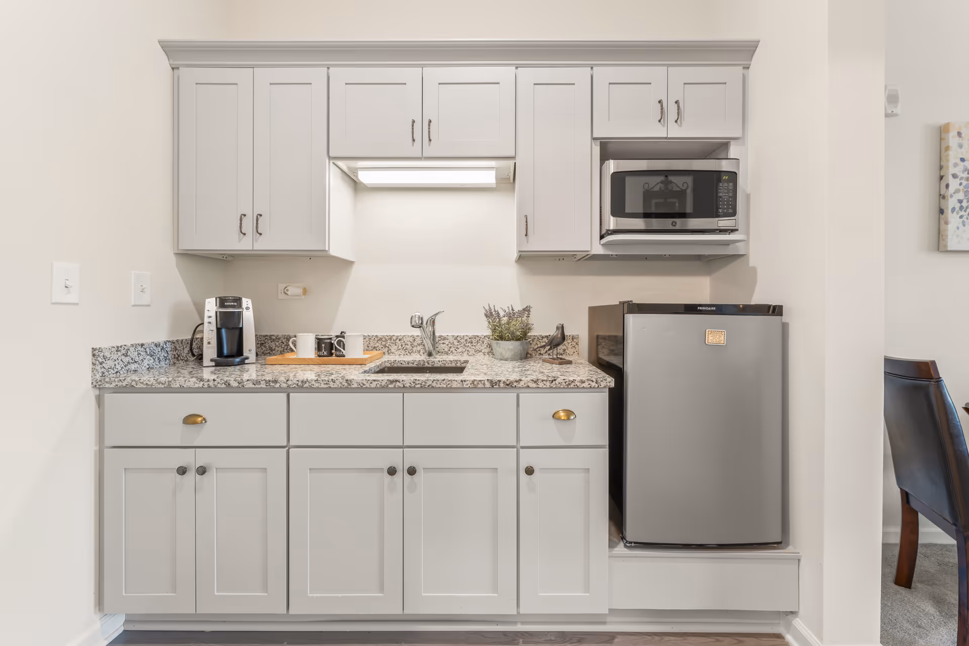 A small kitchen area with light gray cabinets, a granite countertop, a stainless steel sink, a coffee maker, a small potted plant, a decorative bird figurine, a microwave mounted above a compact refrigerator, and a tray with two mugs.