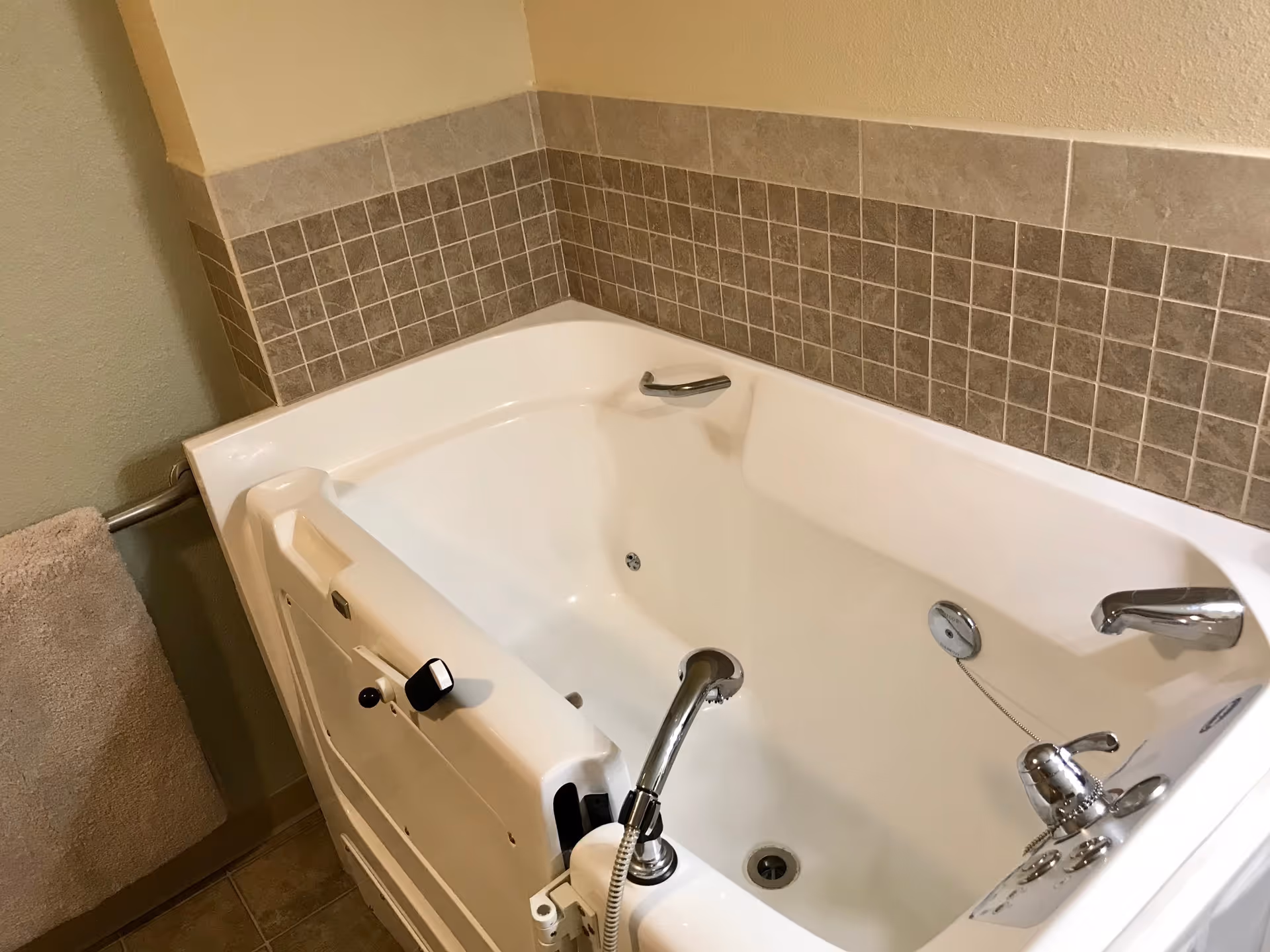 A white walk-in bathtub with a door and safety handle, surrounded by beige and brown tiled walls. A beige towel hangs on a metal towel rack on the left side.