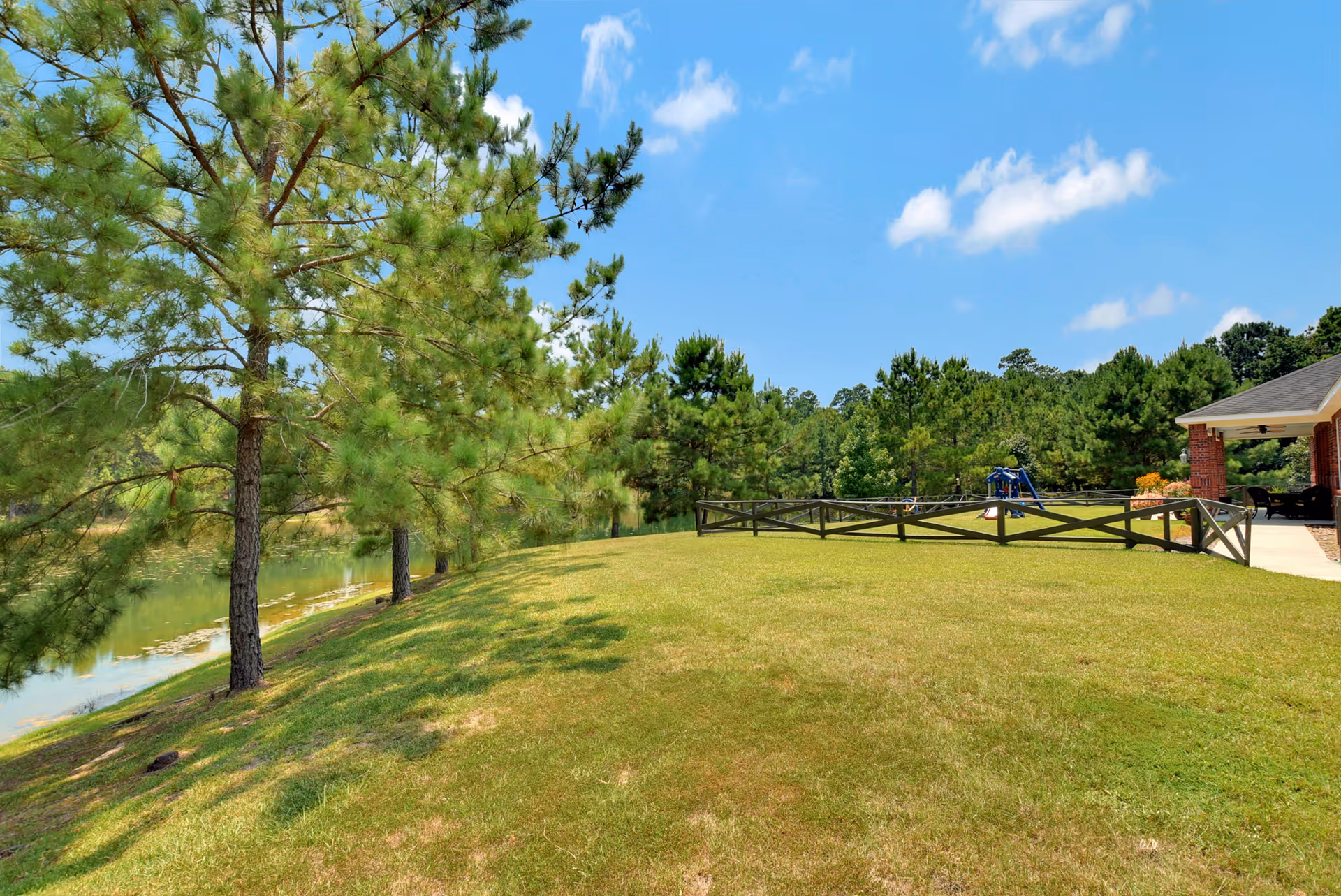 Grassy lawn sloping to a pond lined with pine trees, a wooden fence and a covered porch under a blue sky.