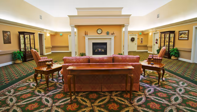 A spacious living room area with a patterned carpet, a central fireplace with a clock and decorative items on the mantel, a red sofa facing the fireplace, two wooden side tables with lamps, and two armchairs. The walls are painted beige with white trim, and there are plants and framed pictures along the walls.