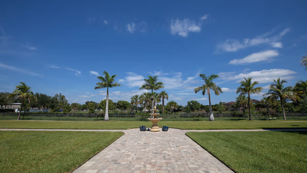 A paved walkway leading to a tiered water fountain in the center of a grassy lawn with palm trees and a clear blue sky in the background.