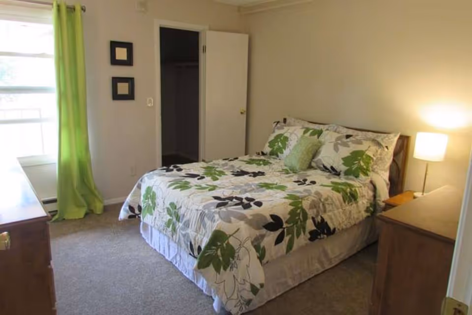 A bedroom with a bed covered in a white comforter with green and black leaf patterns, a green pillow, a wooden nightstand with a lamp, a wooden dresser, a window with green curtains, and an open closet door.