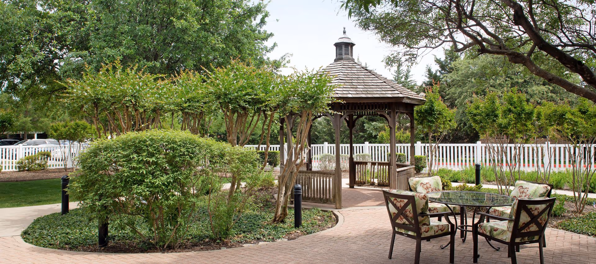 Outdoor garden area with a wooden gazebo surrounded by green trees and bushes. There is a paved walkway leading to the gazebo and a glass-top table with four cushioned chairs featuring floral patterns. A white picket fence and parked cars are visible in the background.