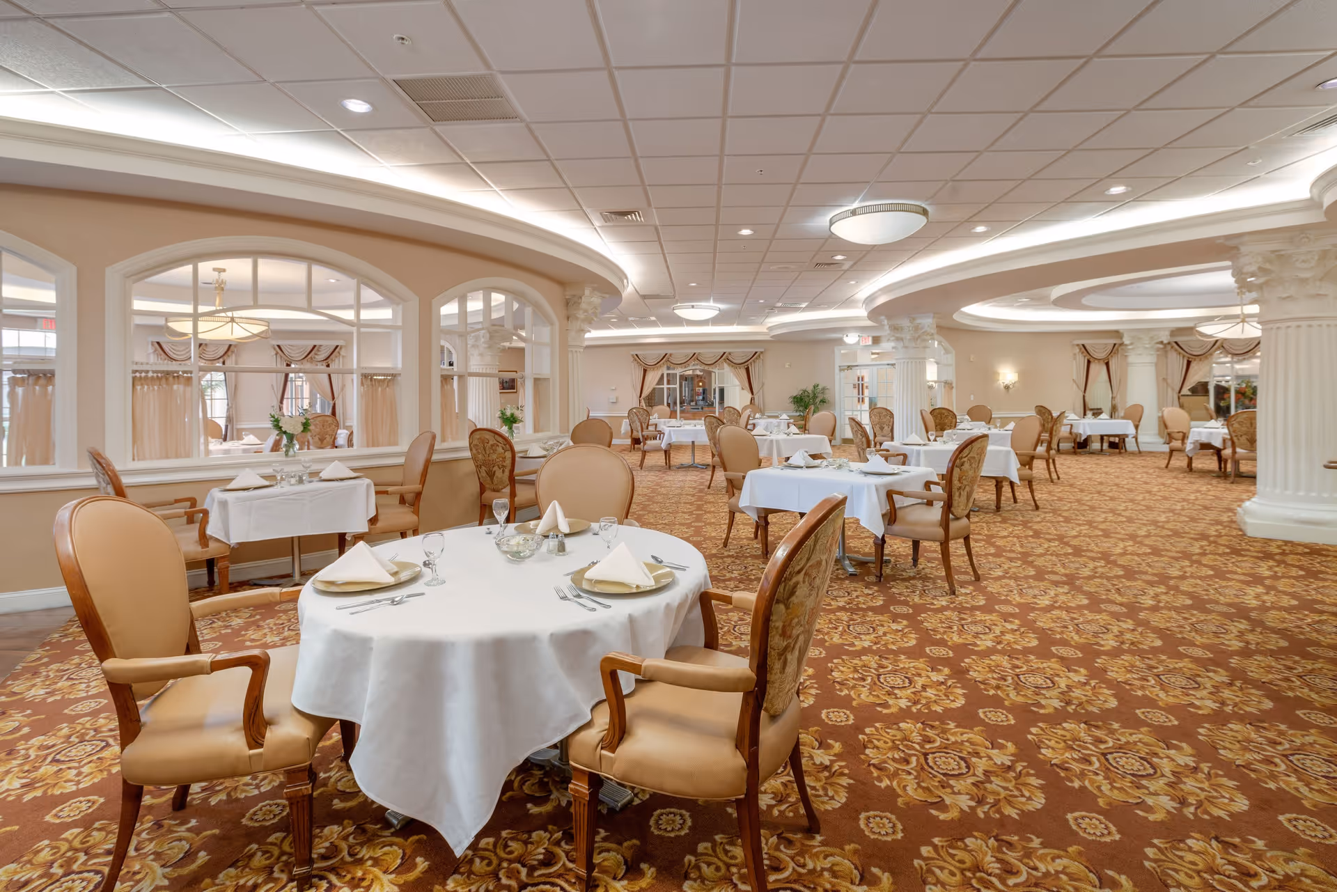 Elegant dining room in a senior living facility with round and rectangular tables covered in white tablecloths, set with napkins, plates, and glassware. The room features ornate carpeting, large columns, soft lighting, and windows with draped curtains.