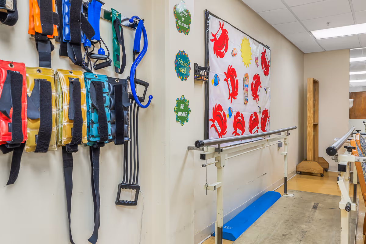 A rehabilitation or physical therapy room with colorful resistance bands hanging on the wall, parallel bars for walking exercises, and a decorative wall hanging featuring red crabs and motivational signs that say 'Love the moment', 'Strive hard', and 'Be brave'.