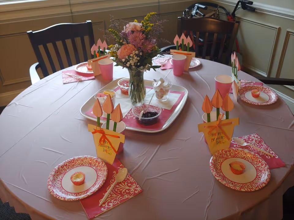 A round table set for a small gathering with a pink tablecloth, decorated with a vase of flowers in the center. Each place setting has a pink and white patterned paper plate with a small cupcake, a pink napkin, a plastic spoon, and a pink cup. There are handmade paper decorations shaped like flower pots with tulips and the words 'Happy Mother's Day' written on them.
