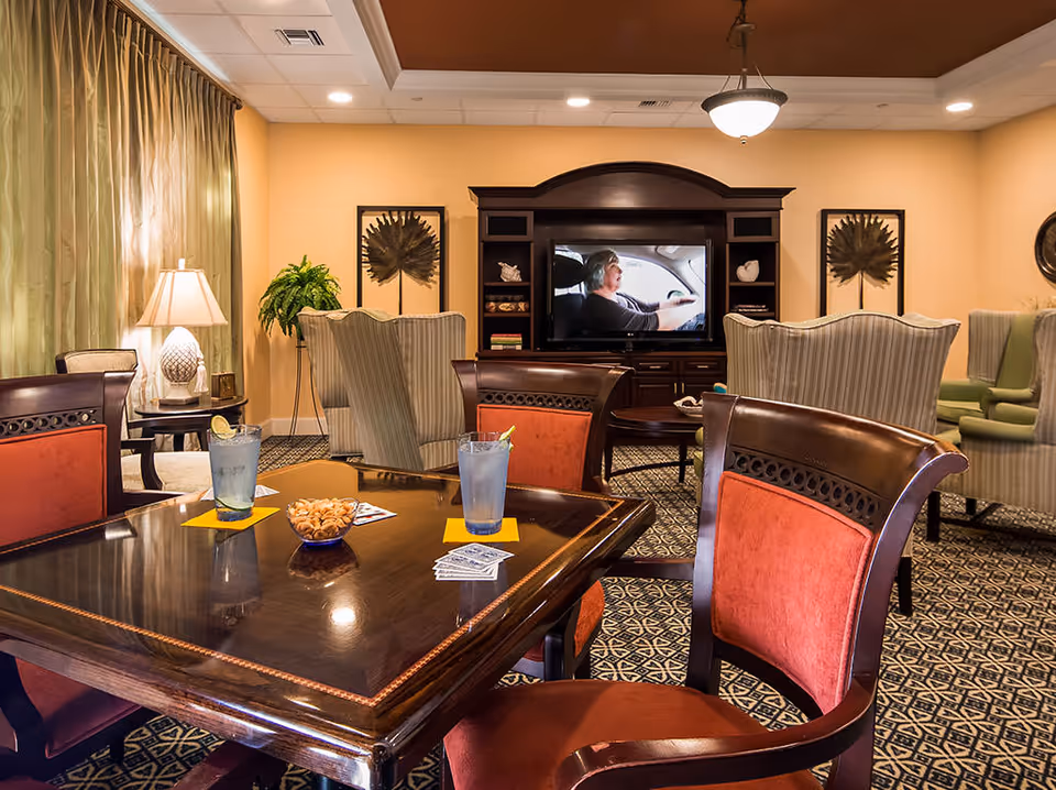 A cozy living room area with a wooden table in the foreground holding two glasses of water, a bowl of nuts, and playing cards. The room features upholstered chairs with red cushions around the table, striped armchairs facing a large flat-screen TV mounted in a dark wooden entertainment center. The walls are painted a warm beige, with framed artwork and a potted plant near a window with green curtains. A ceiling light fixture hangs above.