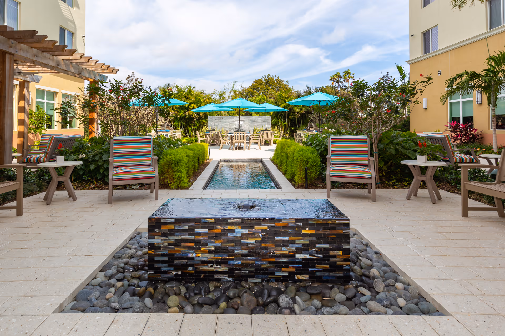 Sunlit outdoor courtyard with a central tiled water feature, a long reflecting pool, striped lounge chairs, and turquoise umbrellas between buildings.
