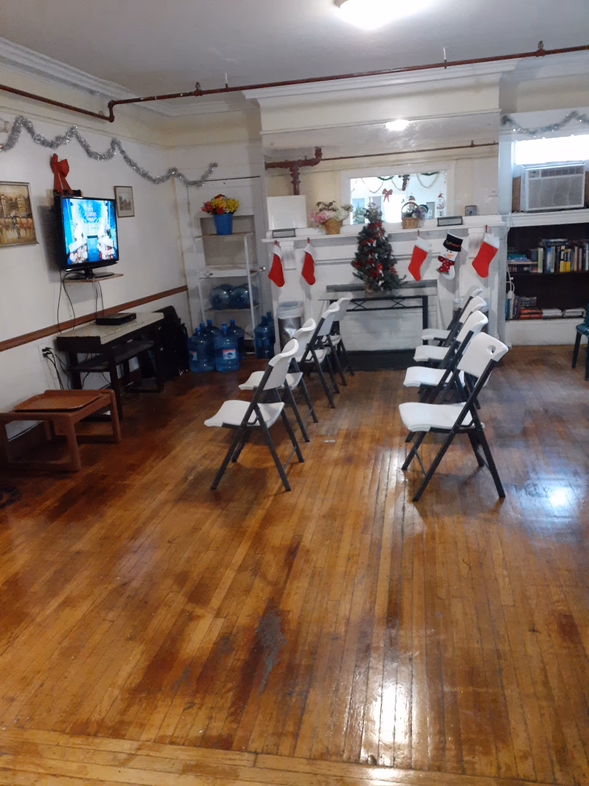 A room with wooden flooring and white folding chairs arranged in two rows facing a TV mounted on the wall. The room is decorated with Christmas stockings hanging on a white mantelpiece with a small Christmas tree and other holiday decorations. There are shelves with water bottles, books, and an air conditioning unit on the right side.