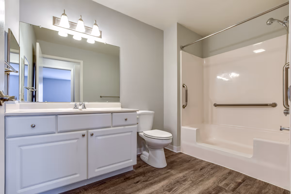 A clean and modern bathroom featuring a white vanity with a large mirror and three light fixtures above it, a white toilet, and a bathtub with built-in grab bars and a shower curtain rod. The floor has wood-like vinyl flooring and the walls are painted light gray.
