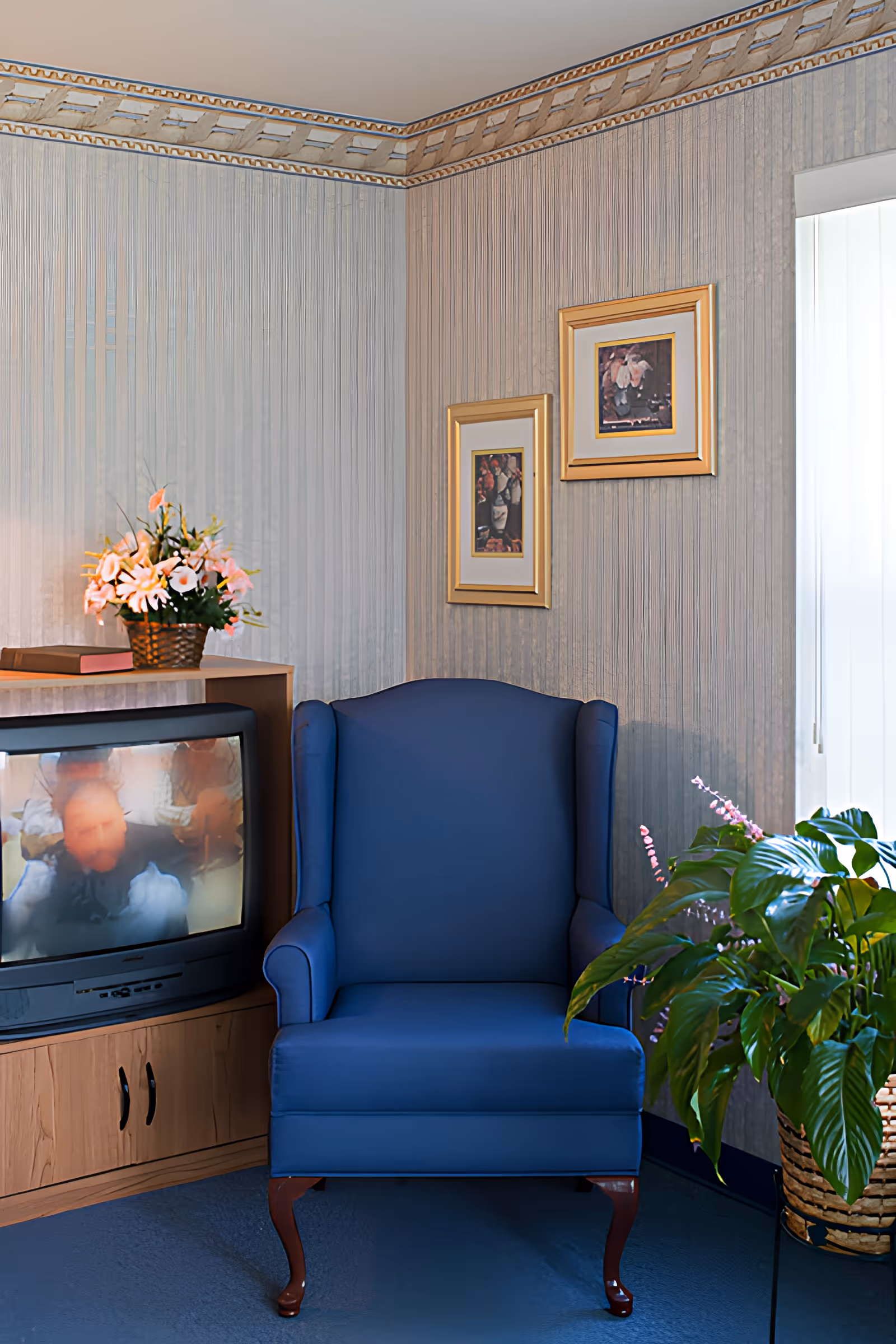 A cozy corner of a room featuring a blue upholstered armchair with wooden legs, a wooden TV stand with a CRT television displaying a blurry image, a basket of flowers on top of the TV stand, two framed floral paintings on the wall, and a large green potted plant near a window with vertical blinds.