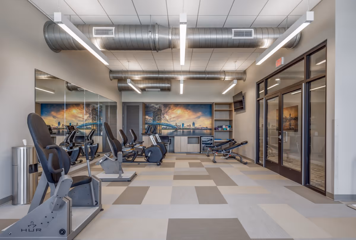 A modern fitness room with exercise bikes and weight benches. The room has a patterned carpet floor, large mirrors on one wall, and a mural of a city bridge at sunset on the far wall. There are exposed ductwork and hanging linear lights on the ceiling, and glass double doors on the right side.