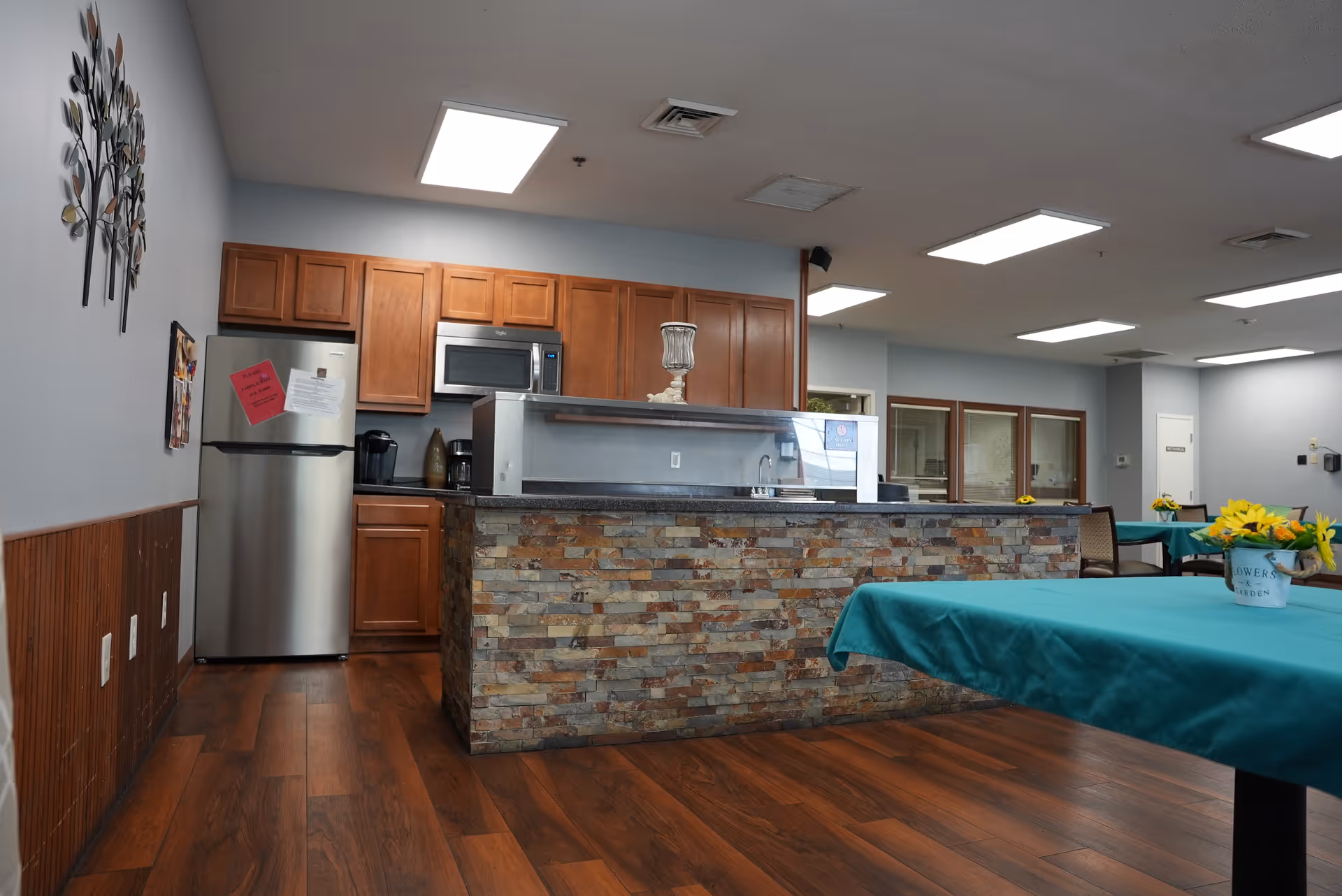 A community dining area showing a stone-front service counter and kitchenette with tables draped in teal tablecloths and vase centerpieces.
