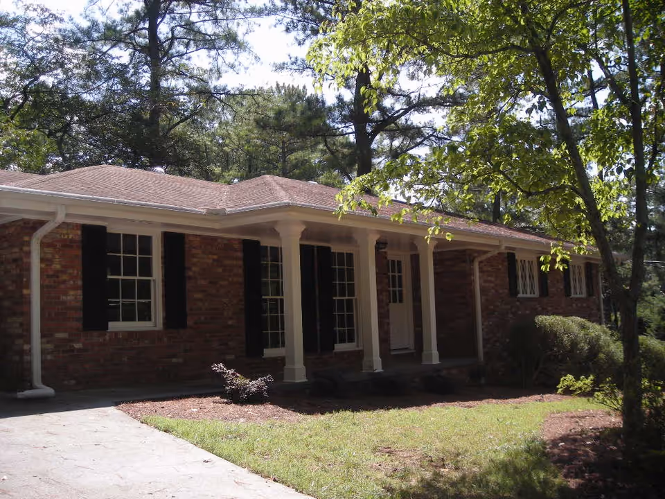 Exterior view of a single-story brick house with white columns supporting a small porch roof. The house has multiple windows with black shutters and is surrounded by trees and shrubs with a grassy lawn in front.