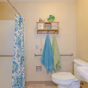 A bathroom with a white toilet, a white sink, a blue and green floral shower curtain, two towels hanging on a wall-mounted rack, and a wooden shelf above the towels holding decorative items and framed pictures.