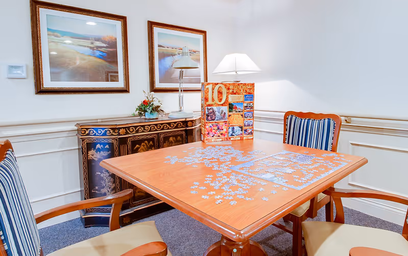 A small room with a wooden table in the center, partially covered with puzzle pieces and a puzzle box. Four wooden chairs with striped cushions surround the table. Against the wall is a decorative black and gold cabinet with floral designs, topped with a small flower arrangement and a silver lamp. Two framed landscape paintings hang on the white wall above the cabinet.