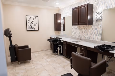Interior view of a salon area with two brown salon chairs, two black hair washing sinks, dark wood cabinets, a large mirror, and a hair dryer chair. The walls are light-colored with a decorative tile backsplash behind the sinks and a framed artwork of hair styling tools.