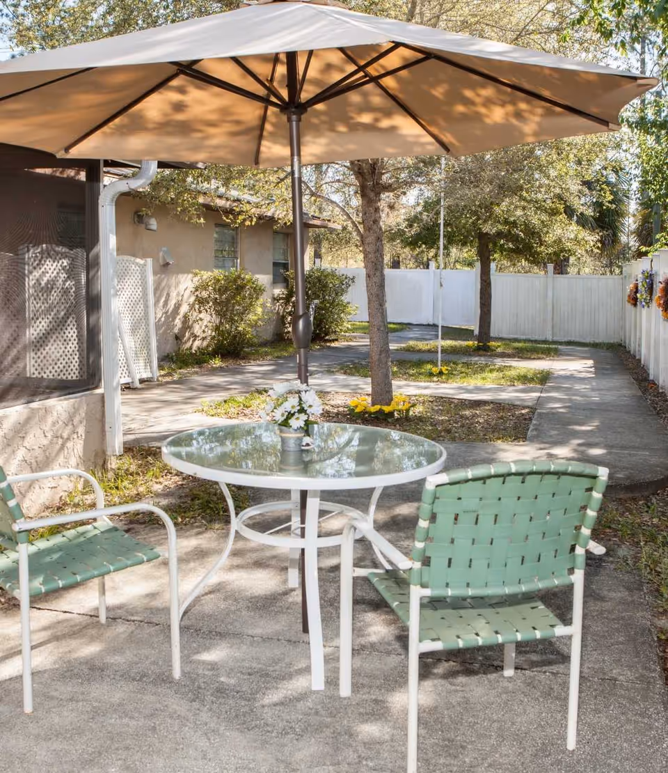 Outdoor patio area with a round glass table and two green woven chairs under a large beige umbrella. The patio is surrounded by trees, bushes, and a white fence with some flowers attached. A building with windows and a downspout is visible on the left side.