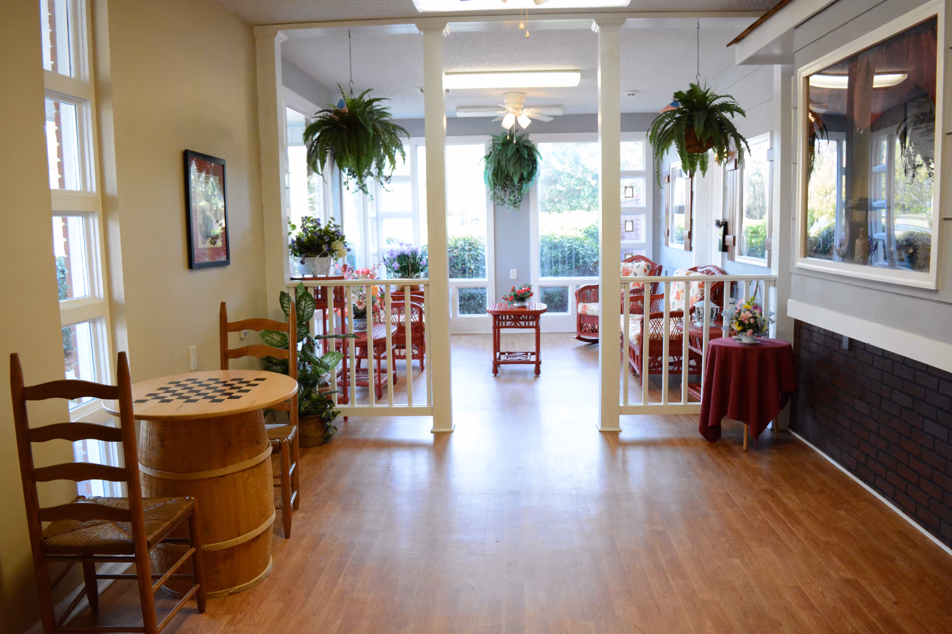 A bright and cozy common area in a senior living facility with wooden flooring, a small round table with a checkerboard pattern and two wooden chairs on the left, hanging green plants, and a seating area with wicker furniture and floral cushions near large windows letting in natural light.