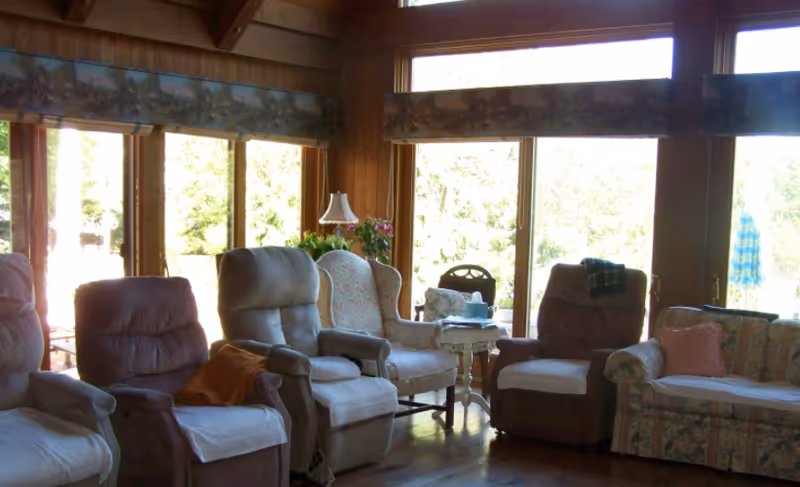 A cozy living room with several cushioned armchairs and a floral-patterned sofa arranged around a small side table with a lamp and flowers. Large windows in the background let in natural light and offer a view of greenery outside.