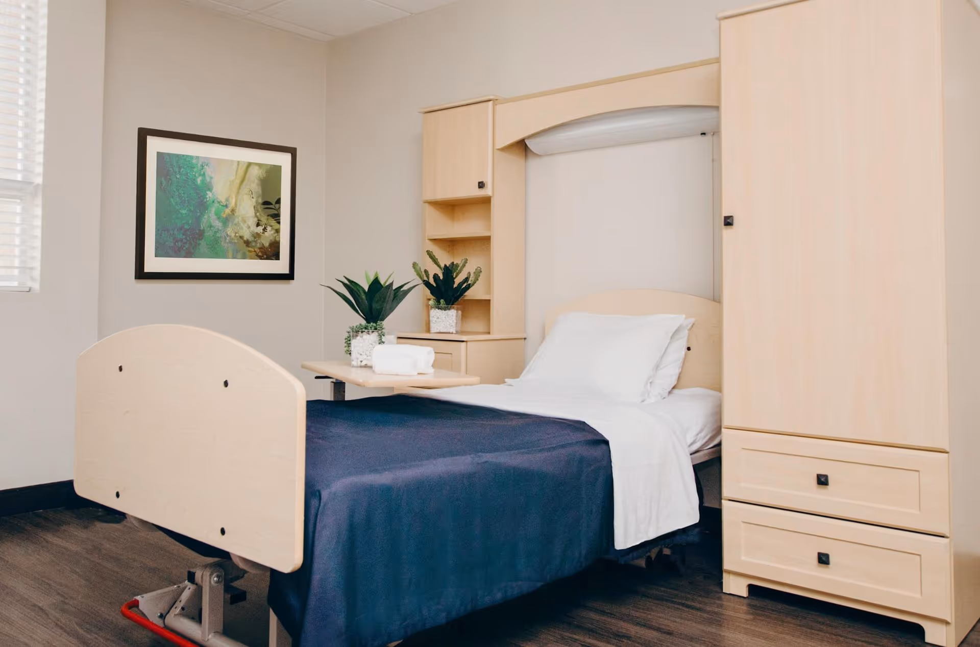 A neatly made single bed with white pillows and a dark blue blanket in a room with light wood furniture including a wardrobe and a built-in headboard with shelves. There are two potted plants on a small table next to the bed and a framed abstract painting on the wall. The room has a window with blinds partially open.