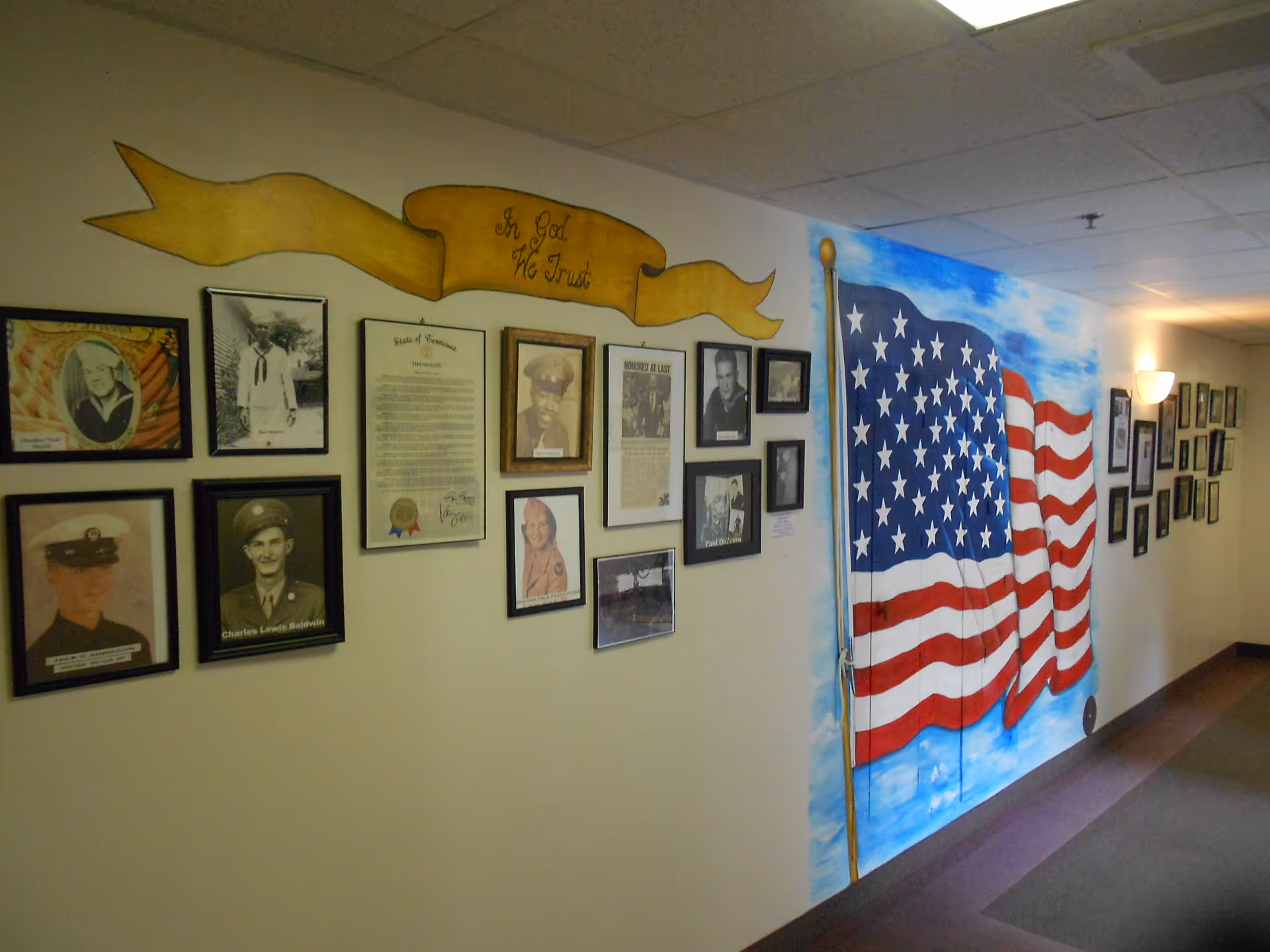 Interior hallway wall decorated with framed photographs and a large painted American flag mural beneath a banner reading 'In God We Trust'.