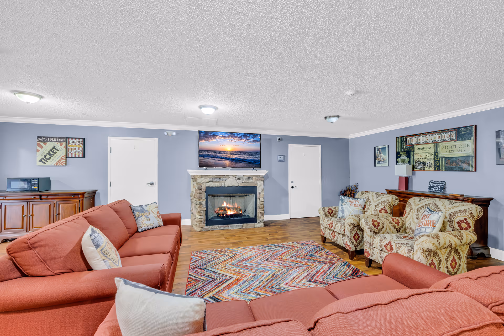 A cozy senior living community lounge with two red sofas and two patterned armchairs arranged around a colorful chevron-patterned rug. A stone fireplace with a TV mounted above it is centered on the far wall. The walls are painted light blue and decorated with framed vintage-style posters. A wooden cabinet with a microwave sits against one wall.