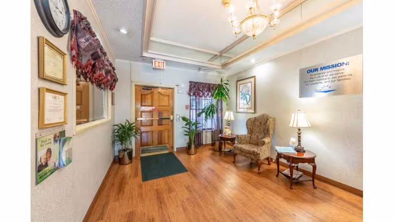 Reception lobby with a wooden exit door, upholstered armchair and side tables, potted plants, and a mission statement sign on the wall.