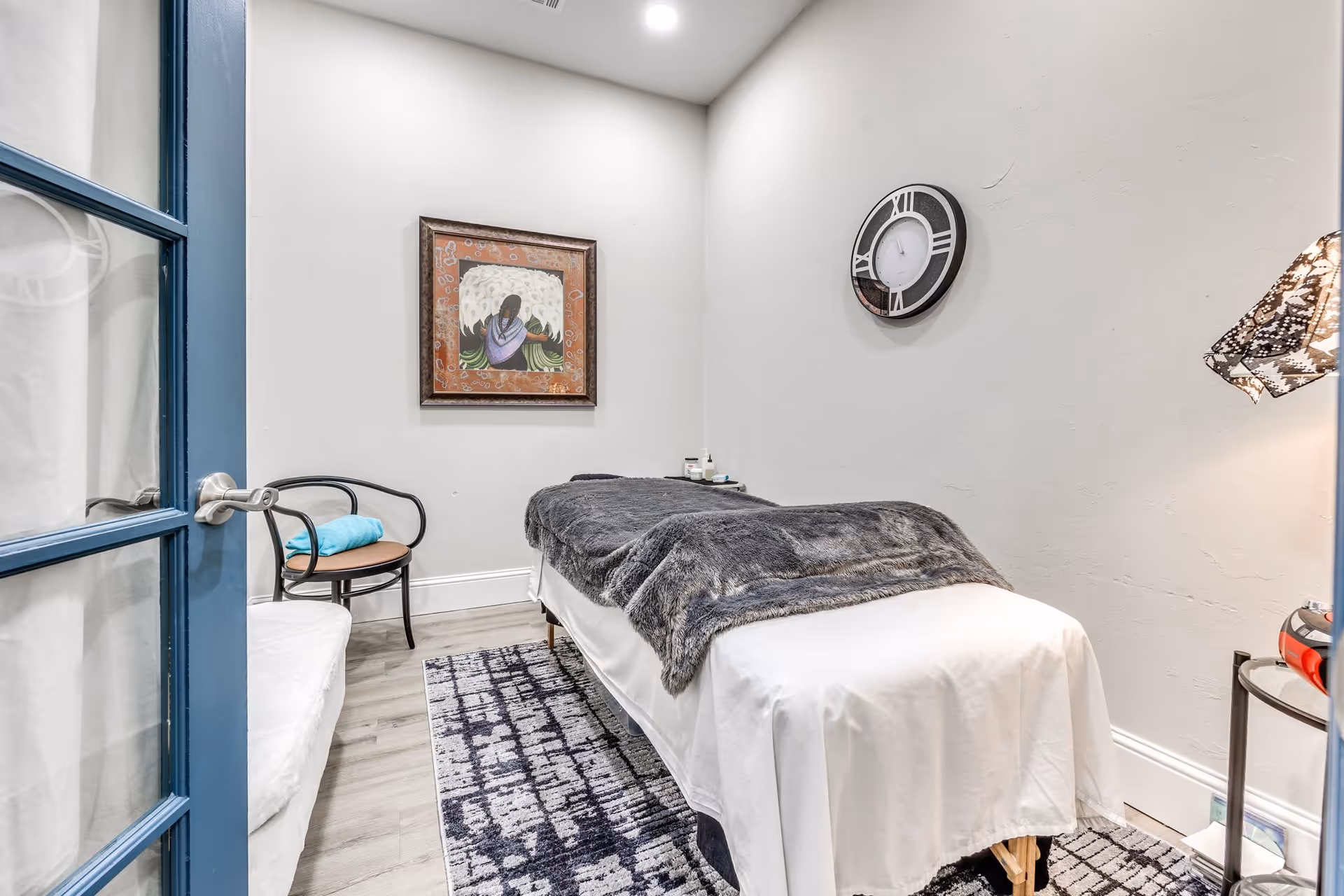 A small, cozy room with a massage table covered in a white sheet and a gray blanket. The room has light-colored walls, a patterned rug on the floor, a framed artwork on the wall, a round wall clock, a chair with a blue towel on it, and a small side table with a lamp and some items.