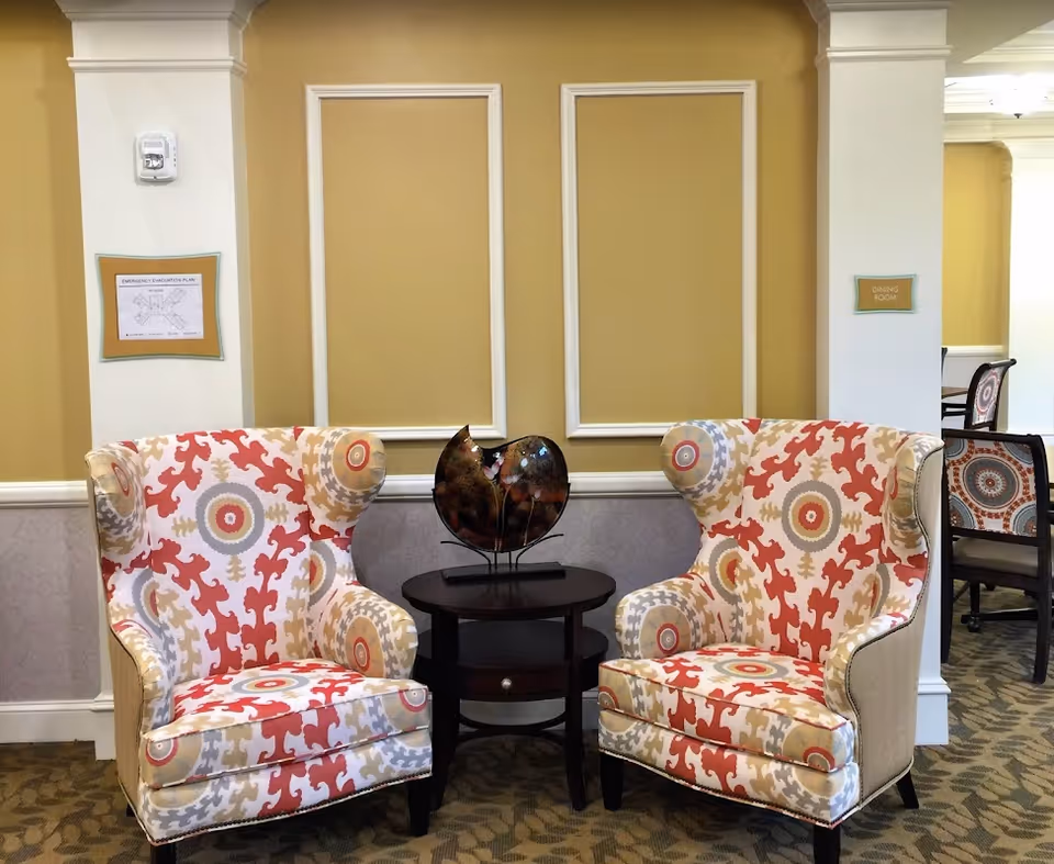 Two patterned wingback chairs with red, beige, and gray designs are positioned on either side of a small dark wooden round table with a decorative sculpture on top. The background features a yellow wall with white trim and two framed signs, one indicating an emergency evacuation plan and the other labeled 'Dining Room'. Part of a dining area with chairs is visible to the right.