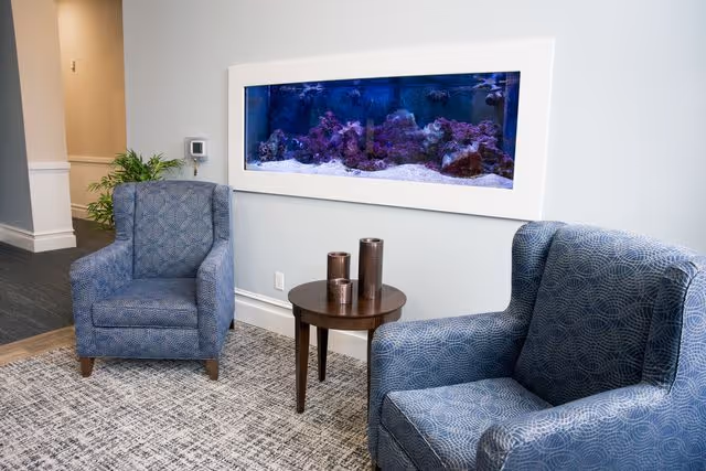 Two blue patterned armchairs positioned around a small round wooden table with three decorative cylindrical objects on it. Behind the chairs, a large rectangular aquarium is mounted on a light gray wall. A plant is visible in the background near a hallway.