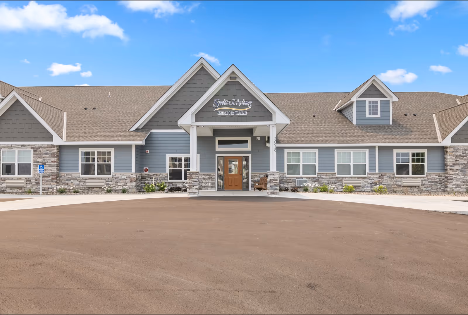 Front exterior view of Suite Living Lakeville senior care facility with a large driveway, stone and blue siding, multiple windows, and a covered entrance under a peaked roof.
