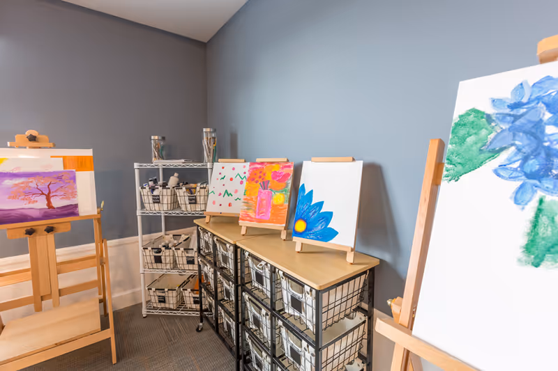 A room with several wooden easels displaying colorful paintings of flowers and a tree. There are storage shelves and baskets filled with art supplies against a gray wall.