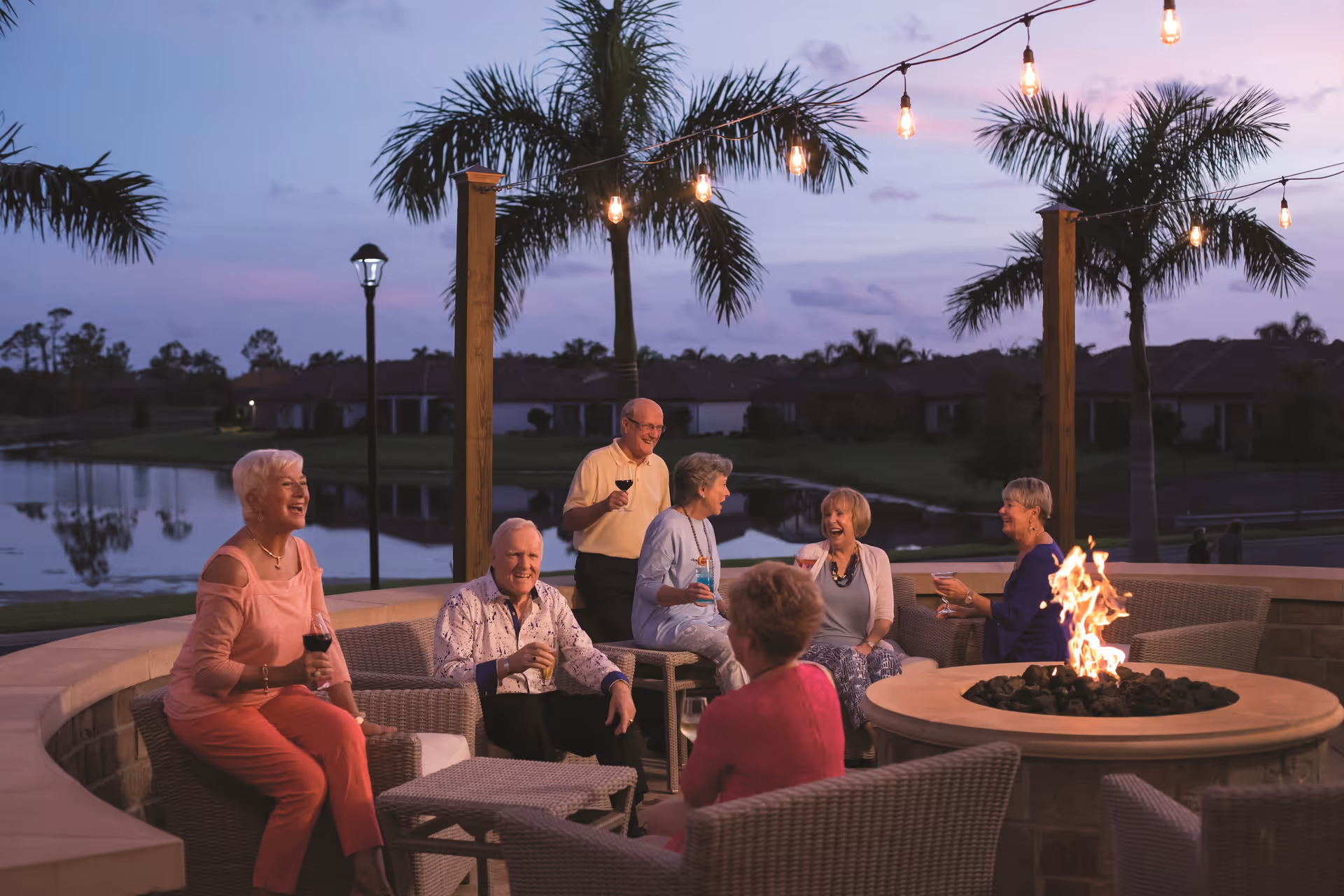 A group of six elderly people sitting and standing around an outdoor fire pit in the evening. They are enjoying drinks and conversation on a patio with string lights overhead, palm trees, and a lake in the background.