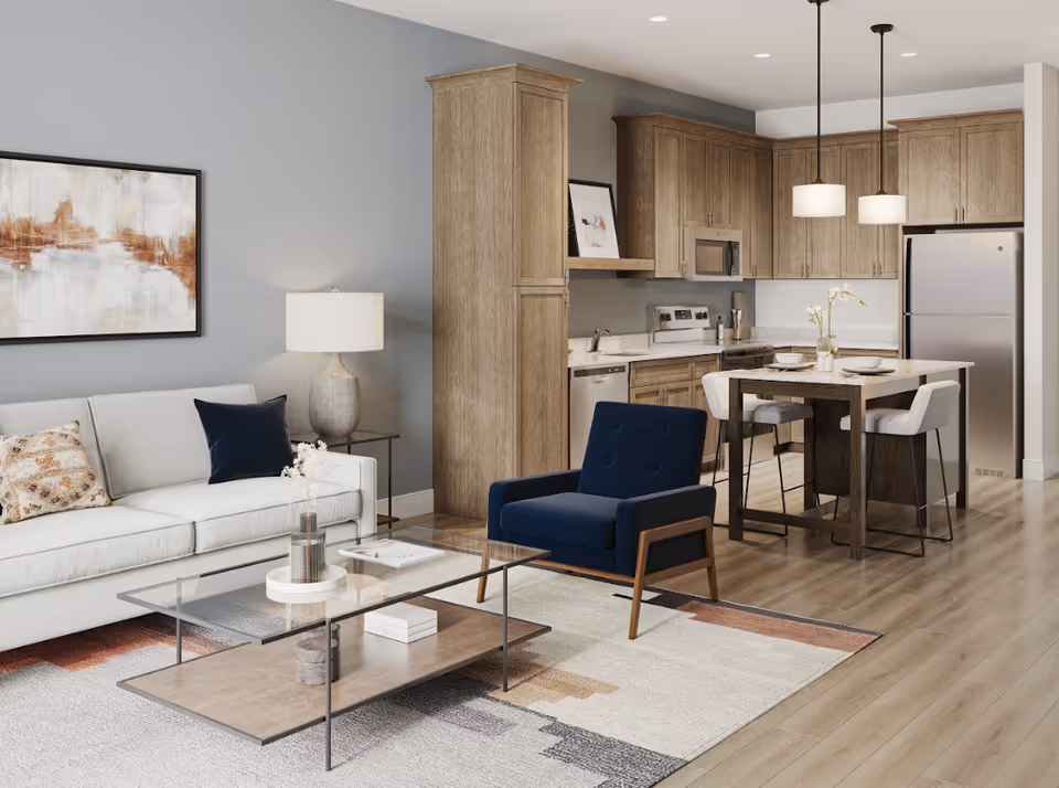 A modern senior living space featuring a light gray sofa with decorative pillows, a navy blue armchair, a glass coffee table on a patterned rug, and a kitchen area with wooden cabinets, stainless steel appliances, and a dining island with two chairs under pendant lights.