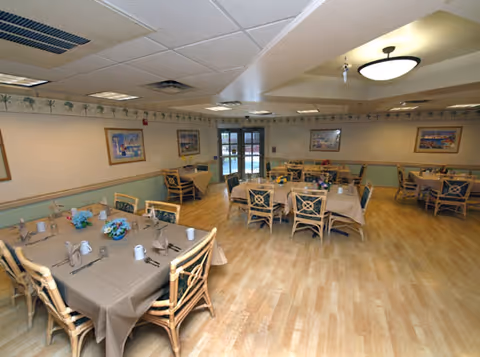 A dining room with several tables covered with beige tablecloths, each set with cups, napkins, and utensils. The room has wooden chairs with cushioned seats, light wood flooring, framed artwork on the walls, and ceiling lights. There is a door with glass panels at the far end of the room.