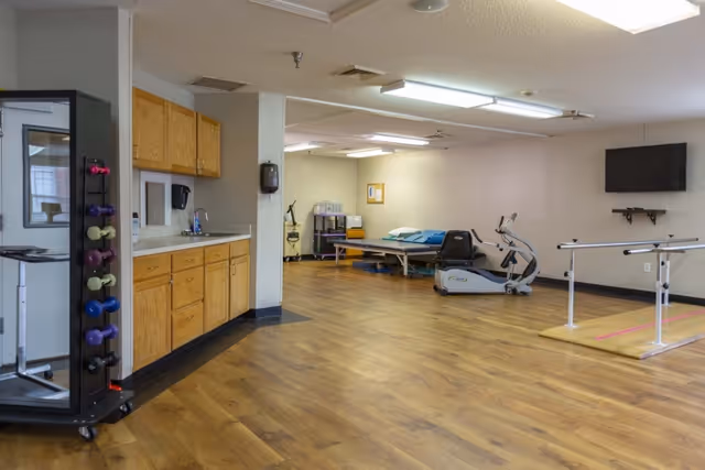 A spacious rehabilitation room with wooden flooring, featuring a stationary exercise bike, parallel bars for walking exercises, a treatment table with pillows, a wall-mounted TV, and a rack holding various colored dumbbells. There are wooden cabinets with a sink on the left side of the room.
