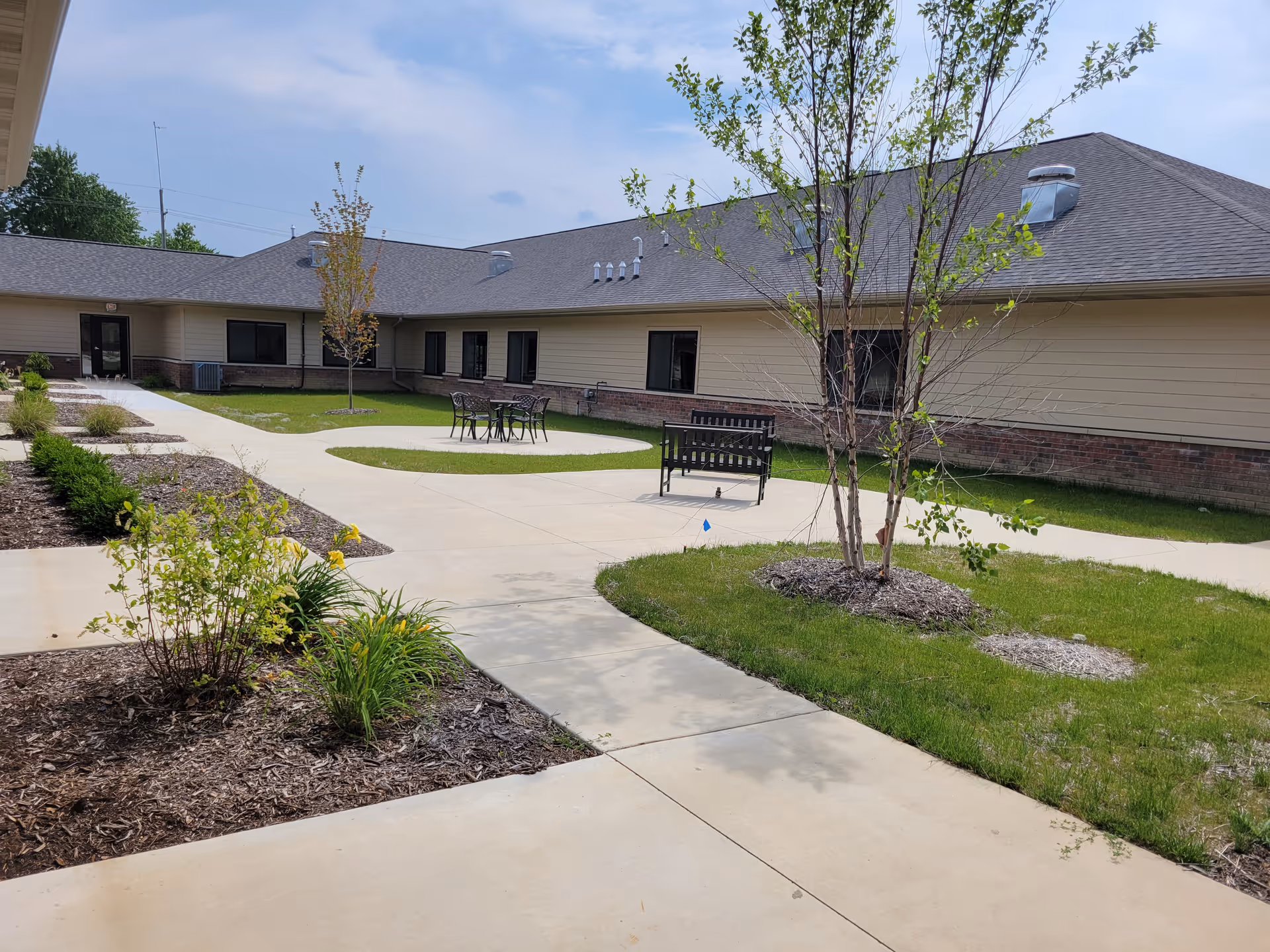 Outdoor courtyard area of a single-story assisted living facility with paved walkways, small trees, shrubs, a bench, and a table with chairs under a partly cloudy sky.