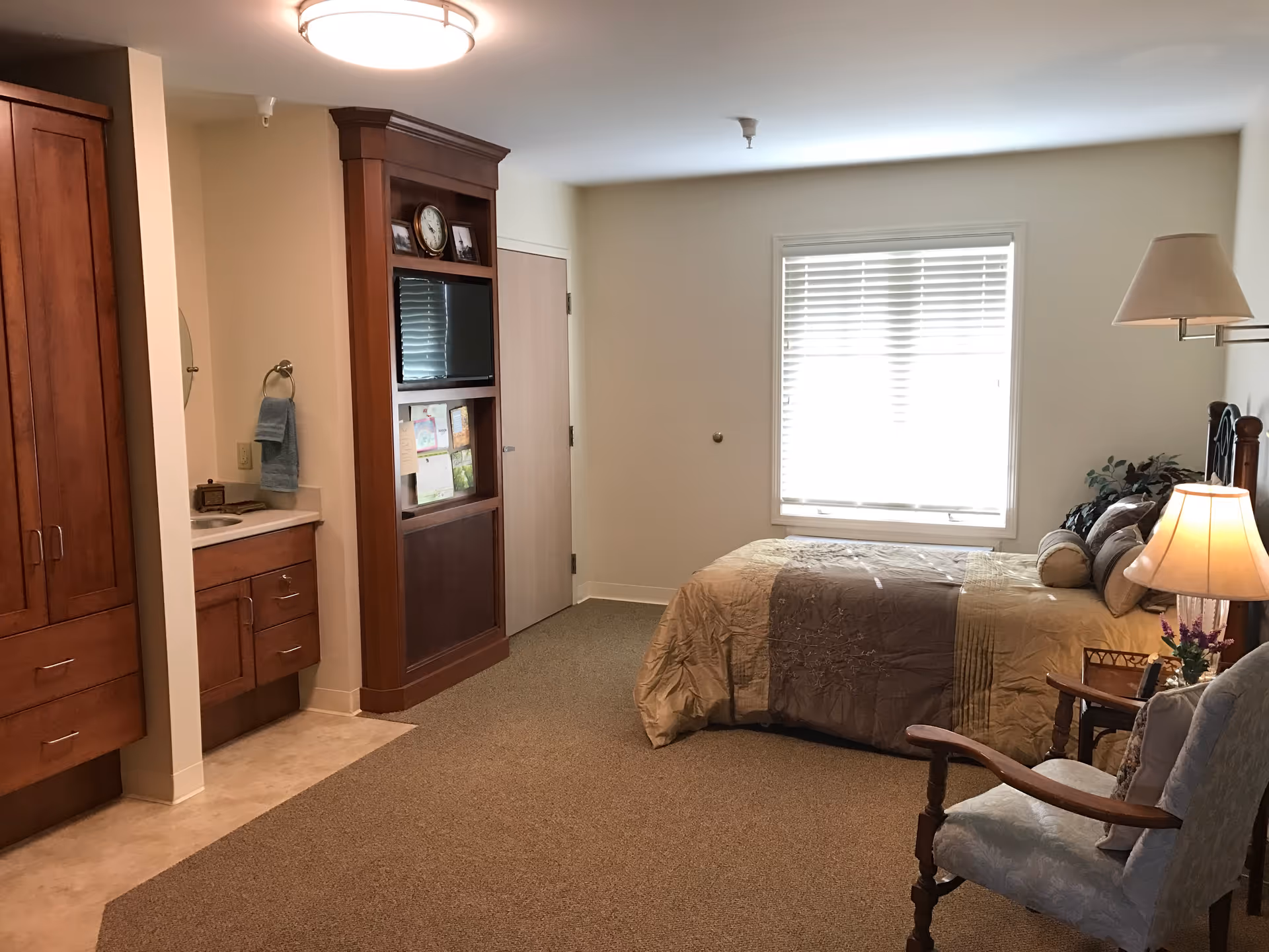 A cozy bedroom in Van Wert Manor featuring a bed with a beige and brown comforter, a wooden armchair with cushions, a bedside table with a lamp and flowers, a window with blinds, a built-in wooden cabinet with a TV and shelves, and a small sink area with a towel hanging nearby.