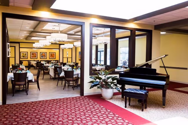 Interior view of a senior living facility showing a dining room with tables and chairs set for meals, visible through large open doorways. In the foreground, there is a black grand piano with a matching bench and a large potted plant next to it. The floor has a patterned red carpet and the walls are painted in warm tones with framed artwork on the dining room walls.