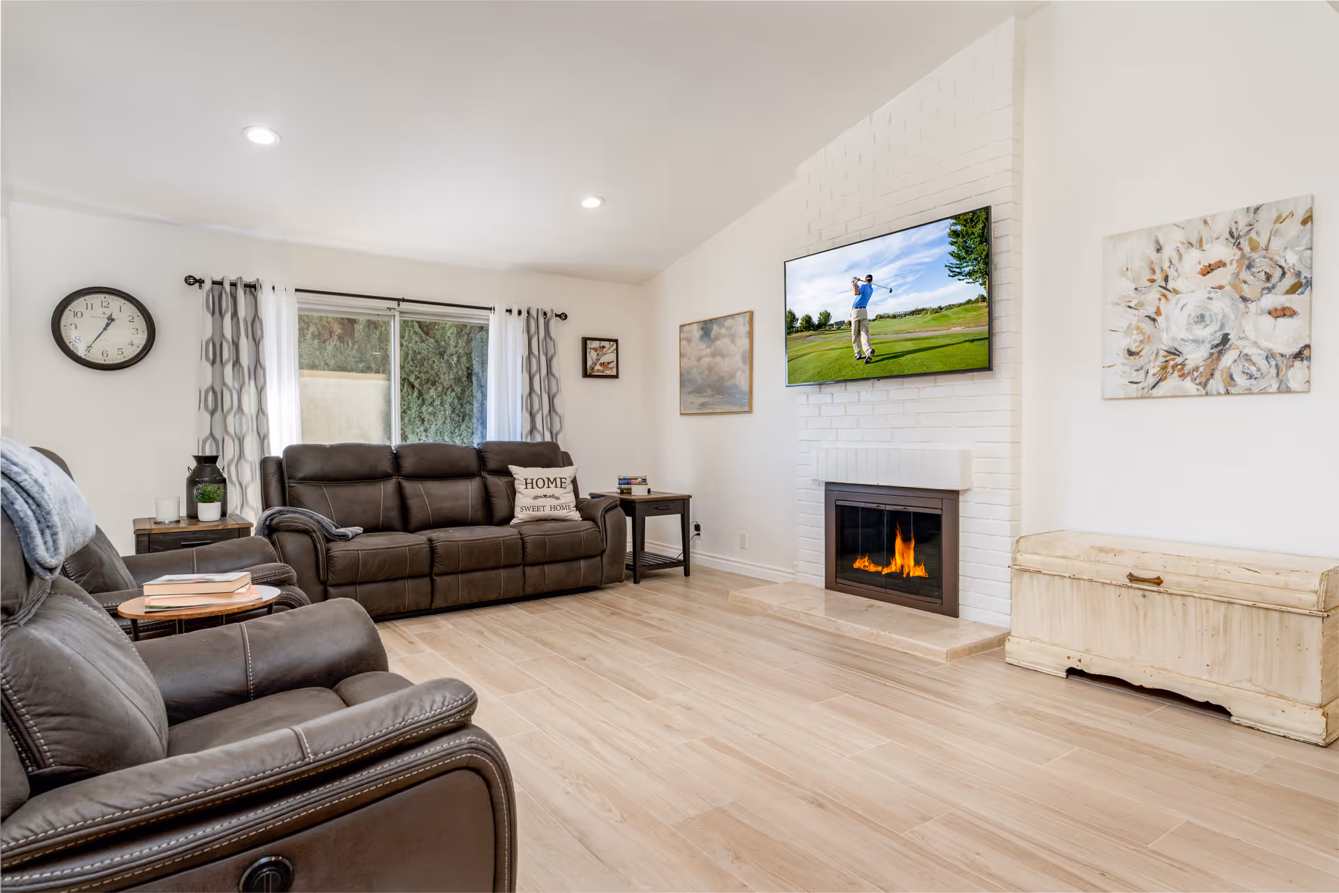 Bright living room with leather recliners and a sofa, a wall-mounted TV above a fireplace, artwork, and wood flooring.