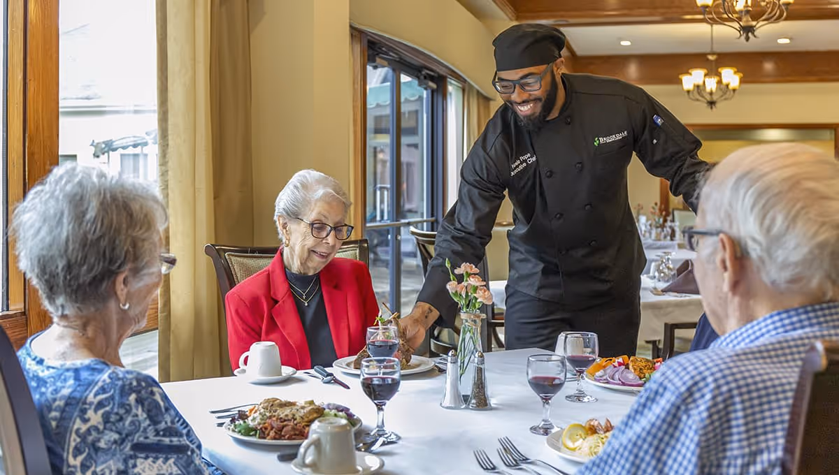 A chef in black uniform and hat serves a meal to three elderly people seated at a dining table in a well-lit dining room with large windows and elegant decor.