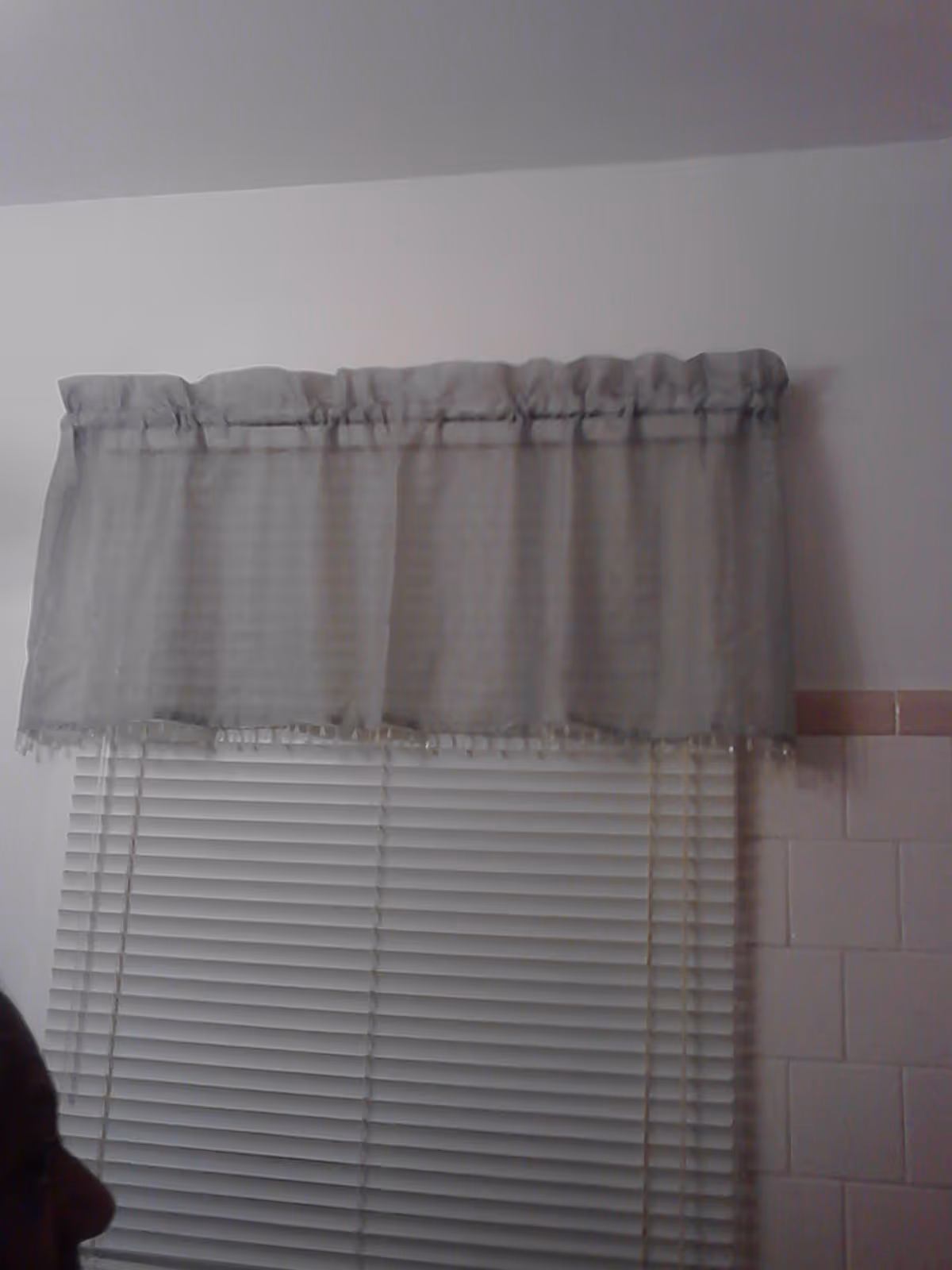 A window with closed white horizontal blinds and a light gray valance curtain above it. The wall is white with a section of light pink tiles on the right side. The silhouette of a person's head is partially visible in the lower left corner.