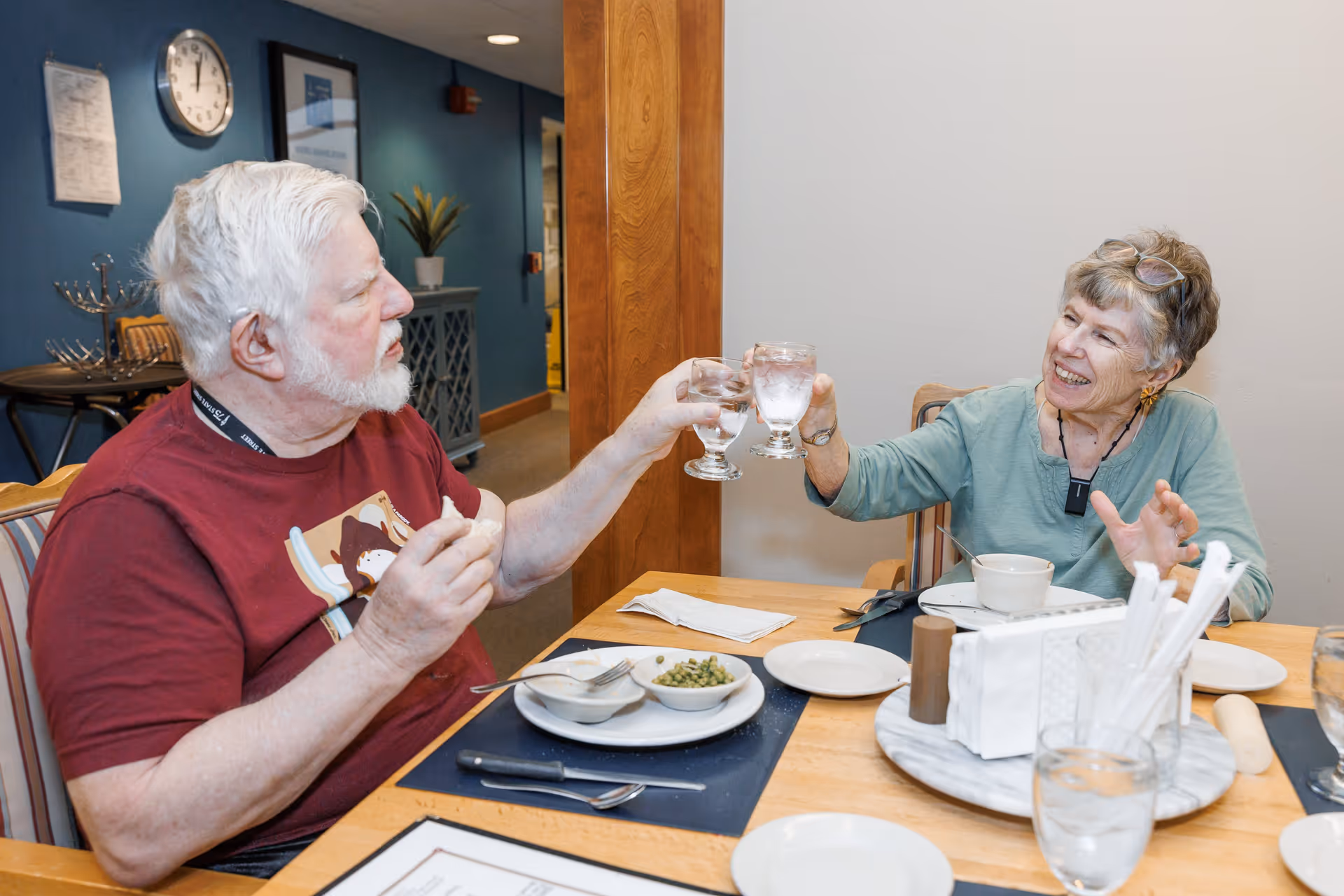 An elderly man and woman sitting at a dining table in a senior living facility, smiling and clinking their glasses of water together. The table has plates with food, utensils, and a napkin holder. The background shows a blue wall with a clock and framed pictures.