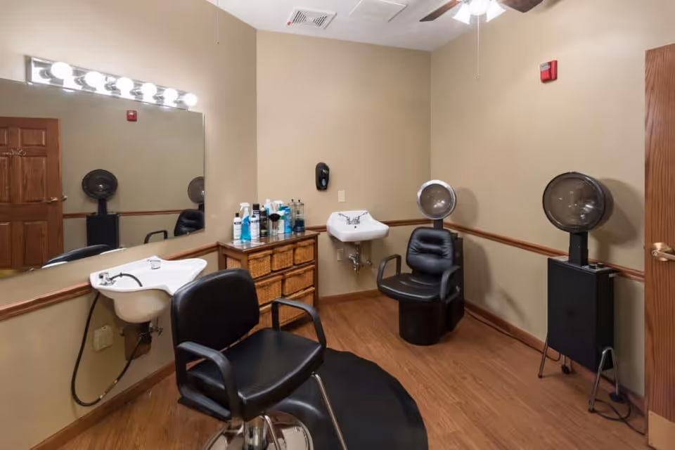 Interior of a small hair salon room with two black salon chairs, a large mirror with lights above it, a sink for washing hair, a cabinet with hair care products, and a hair dryer chair. The room has beige walls and wooden flooring.