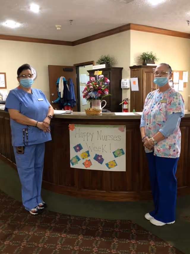 Two healthcare workers wearing masks and scrubs stand on either side of a wooden reception desk with a sign that reads 'Happy Nurses Week!' There is a vase with colorful flowers on the desk, and the background shows a door, coat rack, and cabinets in a well-lit room.