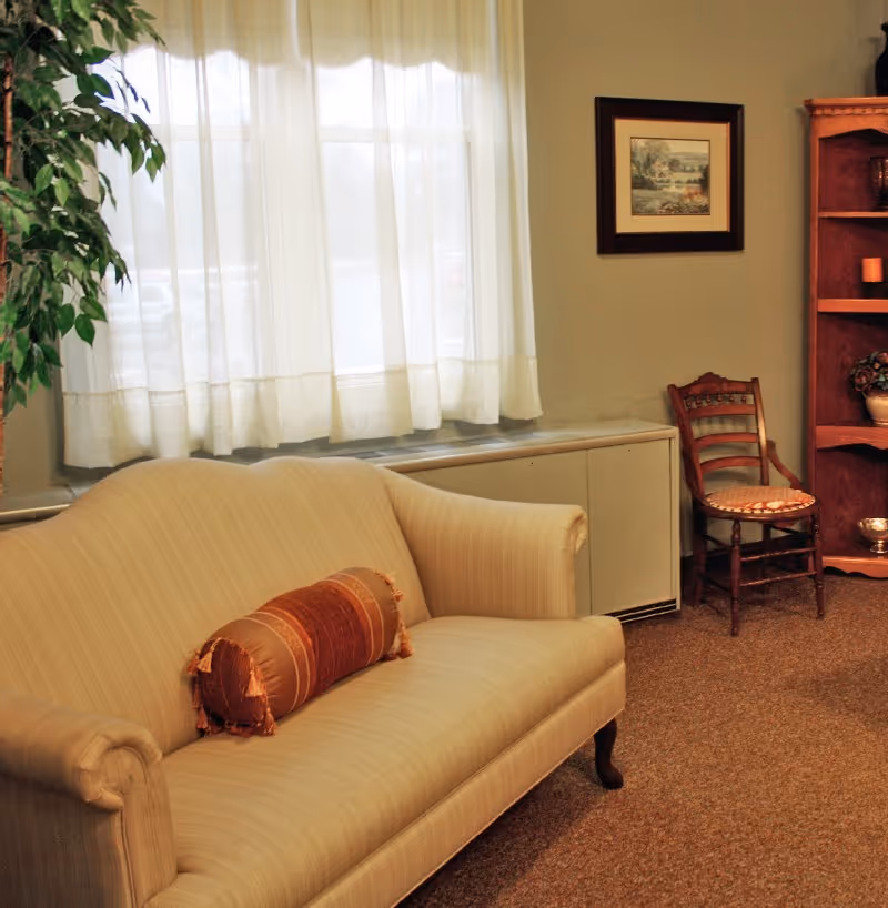 A cozy living room area with a beige upholstered sofa featuring a decorative cylindrical pillow, a wooden chair with a cushioned seat, a wooden bookshelf with various decorative items, a framed landscape painting on the wall, and a window with sheer white curtains letting in natural light.