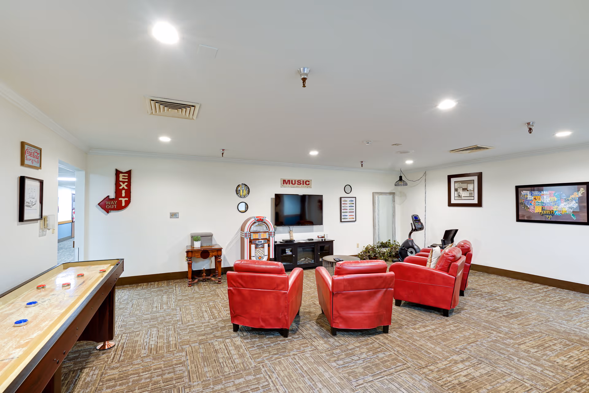 Community lounge with red armchairs facing a TV and jukebox, a shuffleboard table on the left.